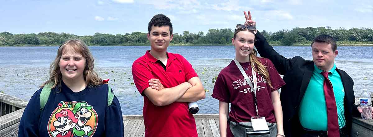a group of 3 special needs adult and one SRACLC staff on a peer by a small lake posing with smile, one of the boys holds up his hand in rabbit ears over the staff members head.