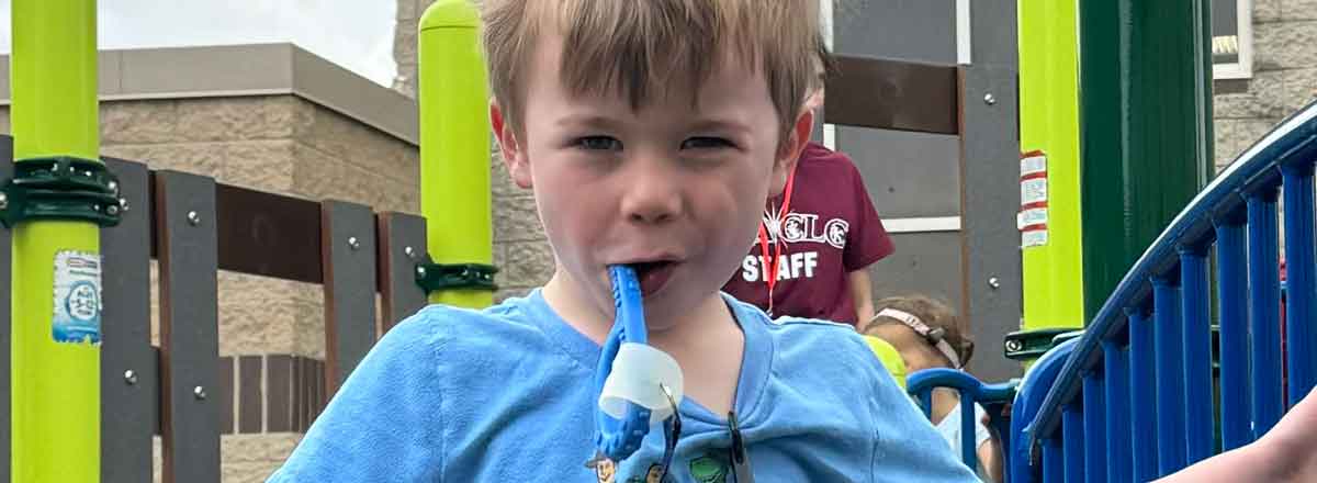A young special needs boy smiling at the camera, his face beaming and fills up teh entire frame, colors of blue and neon green from the playground are in the background.