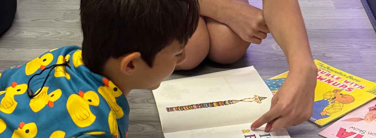 a dark hair special needs boy laying on his tummy reading a book. His shirts has bright yellow ducks on it
