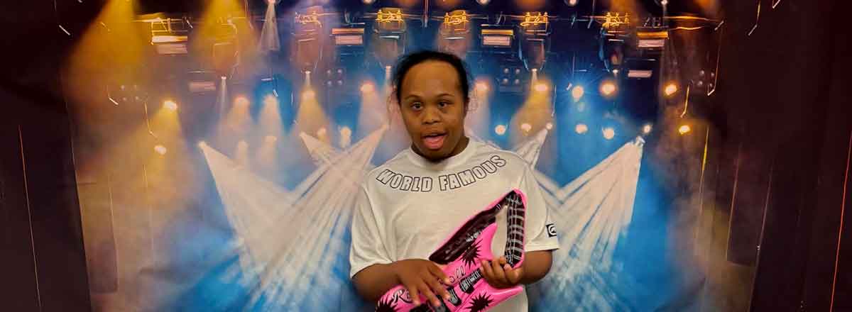 A young special needs man posing in the front of a rock and roll back drop holing an inflatable guitar