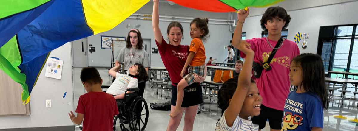a group of younger special needs kids and three staff members playing with a parachute above their heads, the children under smiling and lifting their arms